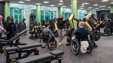 Two wheelchair users get advice from a personal trainer on how to use adaptive equipment in the gym. Other gym goers use benches and treadmills in the background
