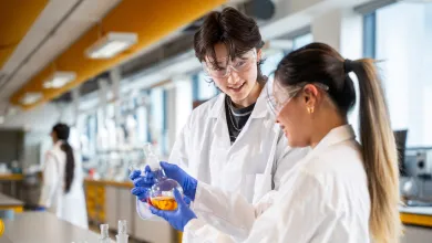 Two students in a laboratory wearing lab coats, safety glasses and gloves hold a glass flask containing orange liquid, with other lab equipment on a bench nearby.