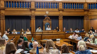 Participants in a formal meeting are seated in a wooden-paneled room with a raised platform at the back where some individuals are discussing.