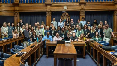 A diverse group of approximately 45 people poses together in a formal setting, featuring ornate wooden panelling and a clock in the background.