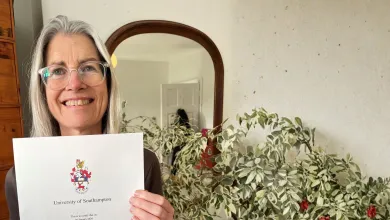 Dr Tara Lamont, with grey hair and glasses, smiles as she holds a University of Southampton certificate, surrounded by a houseplant in a warmly lit room.