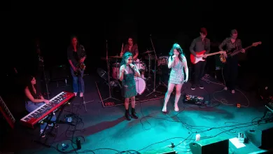 Live band performing on a dimly lit stage. Two vocalists stand at the front with microphones, backed by a drummer, keyboard player, saxophonist, guitarist and bass guitarist.