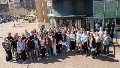 A diverse group of around 50 people stands outside a modern glass building, smiling and posing for a photo on a sunny day.