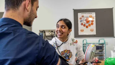 Healthcare trainee measuring a person's blood pressure using an electronic monitor in a clinical training setting.