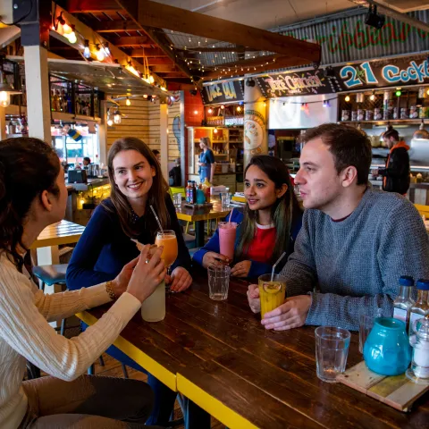 Group of students sat at table in a restaurant bar, talking and laughing over drinks.