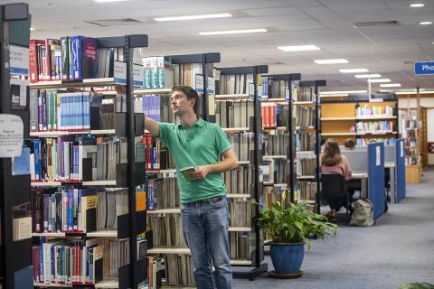 A student selects a book from a shelf while other students work at desks in the background