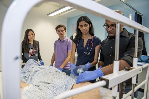 A group of students watch as one of them works on a dummy patient in a hospital bed
