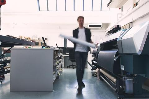 A student walking between several industrial-sized printers and a long work bench lined with guillotine paper cutters