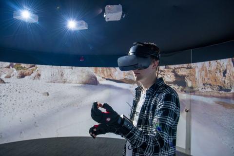A student wearing a virtual reality headset and gloves, standing in front of a 360 degree video wall displaying what he can see.