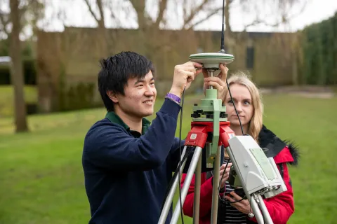 Two students conduct an experiment outdoors using a large piece of survey equipment on a tripod.