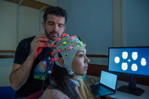 In a dark room, a man attaches electrodes to an EEG cap worn by a seated student. The cap is attached to a computer behind the pair, which displays brain activity data.