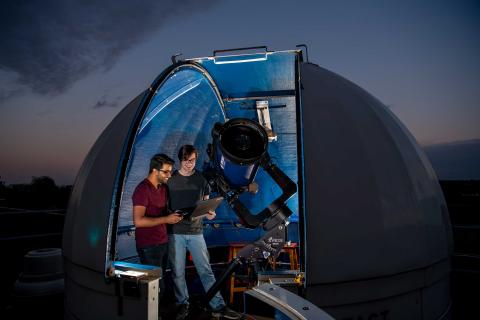 A small, domed rooftop observatory opens onto a starry night sky. Inside the observatory, two students use a telescope and a laptop to collect data.