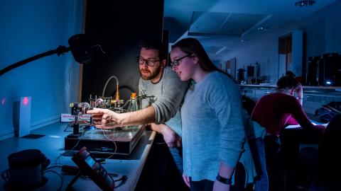 Two students set up a small experiment with a laser, in a darkened laboratory, surrounded by technical equipment.