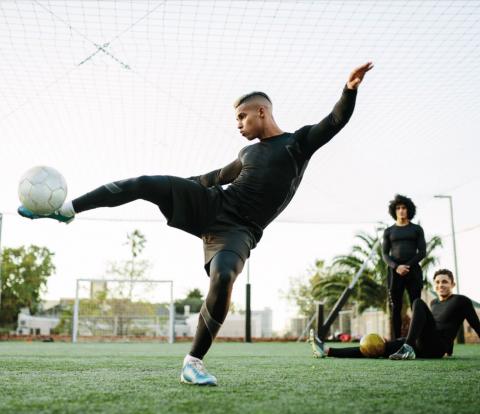 A young male footballer raising his leg parallel to the ground, to kick a ball towards the camera.