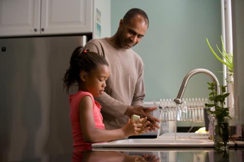 A father smiling down at his young daughter as she washes her hands carefully with soap.