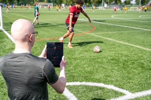 Dr Devon Lewis standing by a football pitch as a player kicks the ball. Devon is holding an iPad that shows live data streamed from a wearable sensor.