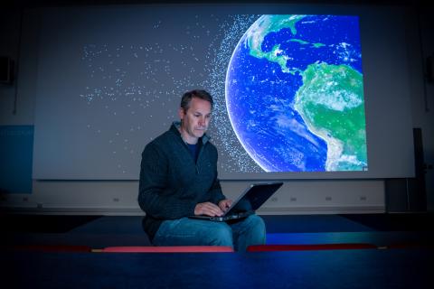 Professor Hugh Lewis sitting on the front bench of a lecture theatre as he consults his laptop. A graphic of satellites orbiting Earth is projected on the screen behind him.