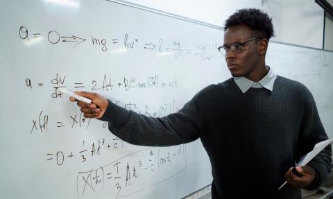 A maths teacher writes on a white board and looks intently at the equations. We imagine he is in front of a classroom of pupils.