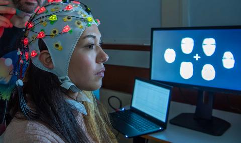 A study participant wears an EEG cap and is seated close to a screen displaying data.