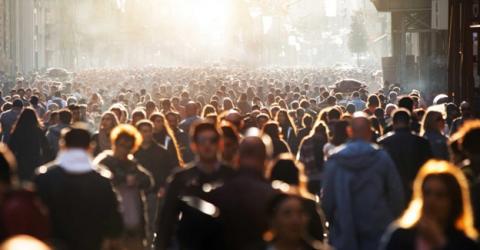 A large crowd of people stretches as far as the eye can see. A low, bright sun highlights a number of individuals who make up the crowd