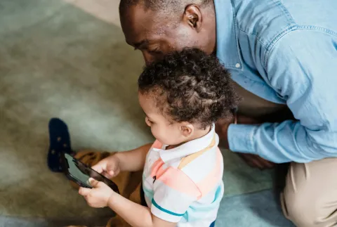 Young child playing on a phone as an adult watches them over their shoulder looks 