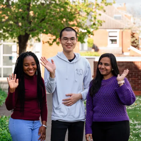 Three students stood outside smiling and waving at the camera.