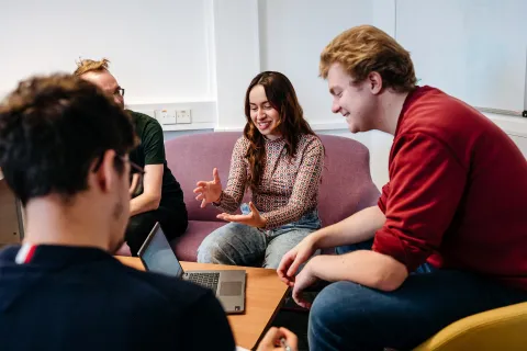 Four people sitting around a small table in a casual meeting setting. One person is speaking and gesturing with hands, while others are listening attentively. A laptop is open on the table, and the group is seated on cushioned chairs in a bright room with a whiteboard in the background