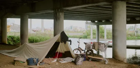 A makeshift shelter set up under a bridge near a river, with personal belongings, a bicycle, and a shopping trolley. The image reflects urban homelessness and temporary living conditions