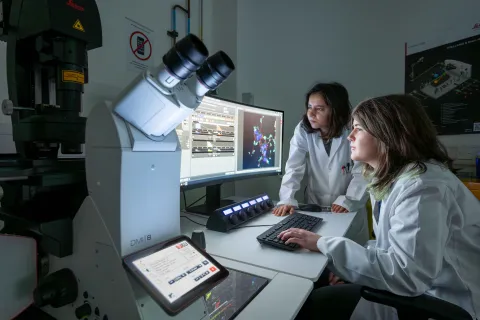 Two students in white lab coats using the microscopy lab facility. They're both analysing data on a computer monitor and there's a large microscope to the side of them.