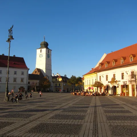 Small, Romanian market square with a clock tower, cobbled paving and painted white buildings, blue sky overhead.