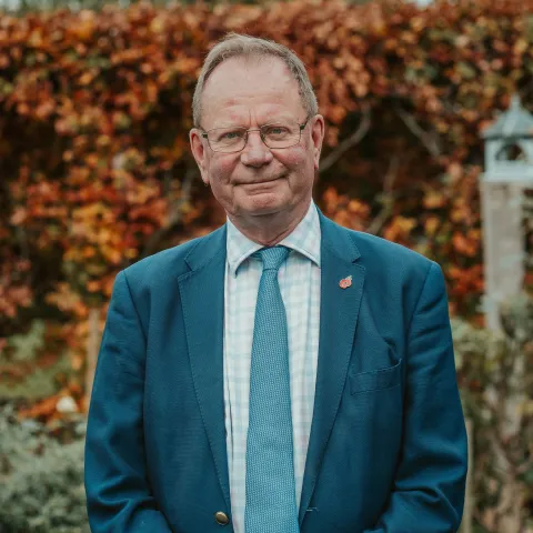 Portrait of Professor Stephen Holgate in a blue suit standing outdoors in front of autumn foliage.