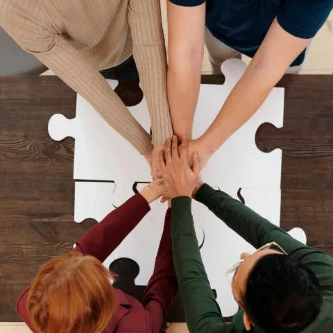 Hands of four people stacked over a white puzzle piece on a wooden table.