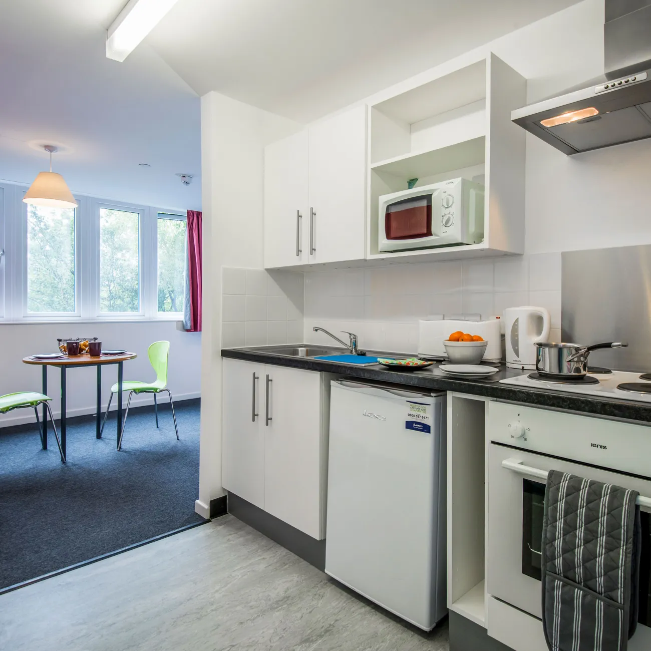An open plan kitchen area with laminate flooring and modern white kitchen units. A small table, chairs and open window are visible in the background.