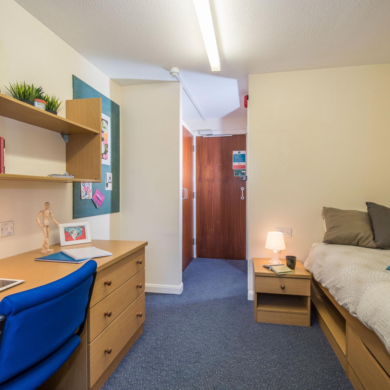 A student bedroom containing a working desk area and a neatly made bed. In the background a door opens to a bathroom.