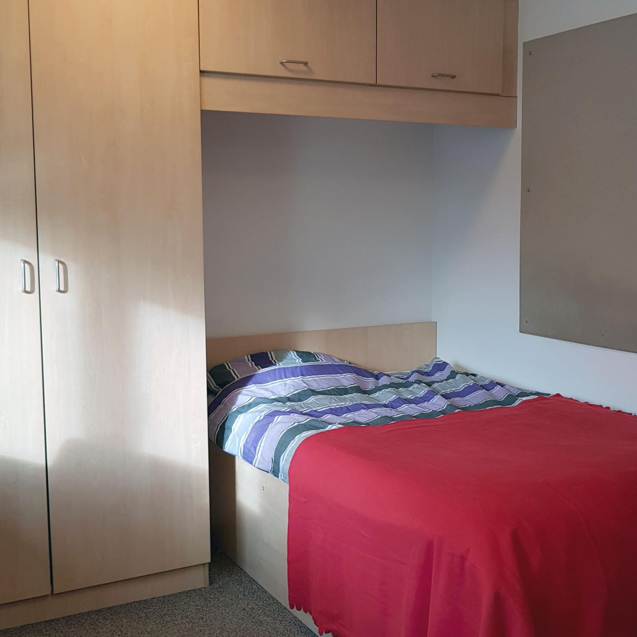 A student bedroom showing large double bed with red bed sheets, full height wooden wardrobes and cupboards. Light streams in through an unseen window.