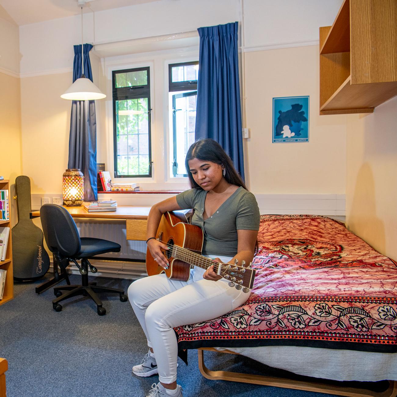 A student plays a guitar while sitting on a bed in a bedroom. In the background, shelves and a desk with books can be seen.