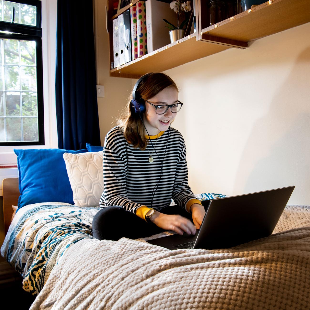 A student sits cross-legged on a bed, working on a laptop. The room is partially lit with a lamp, a window lets in some light.