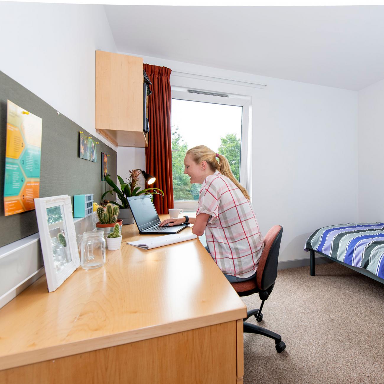 A spacious, bright student bedroom showing a single bed, desk and open window. A student sits at the desk, working on a laptop.