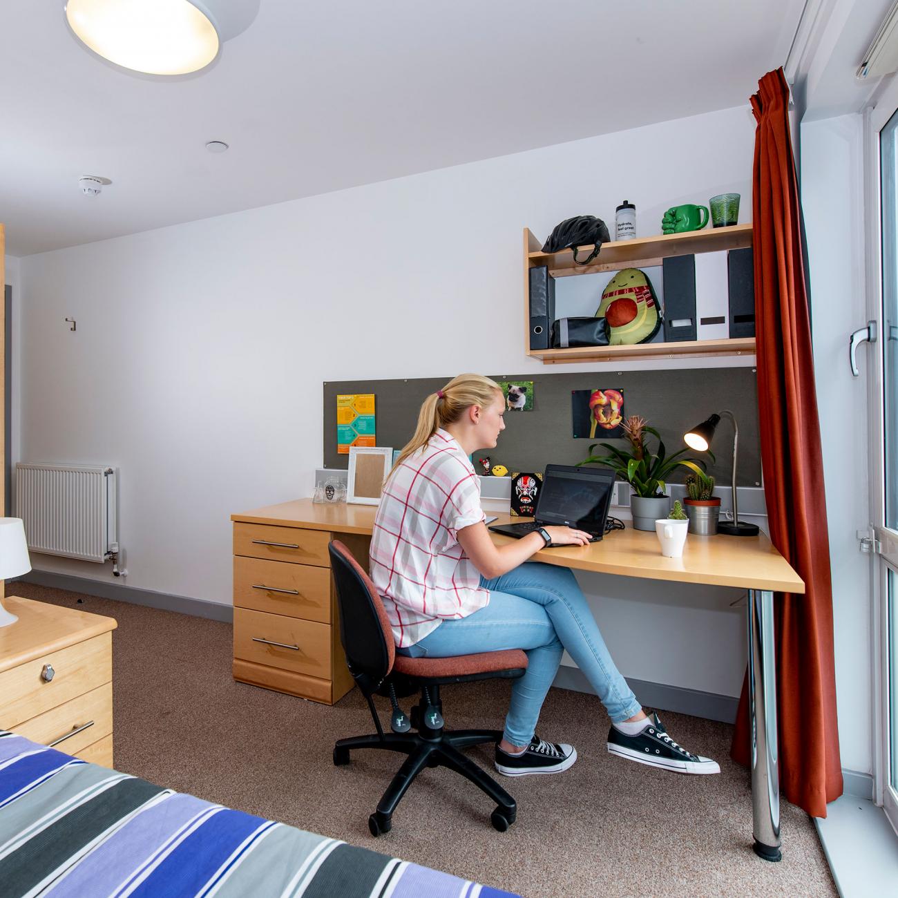 A spacious student bedroom showing a single bed, desk and wardrobe. A student sits at the desk, working on a laptop.