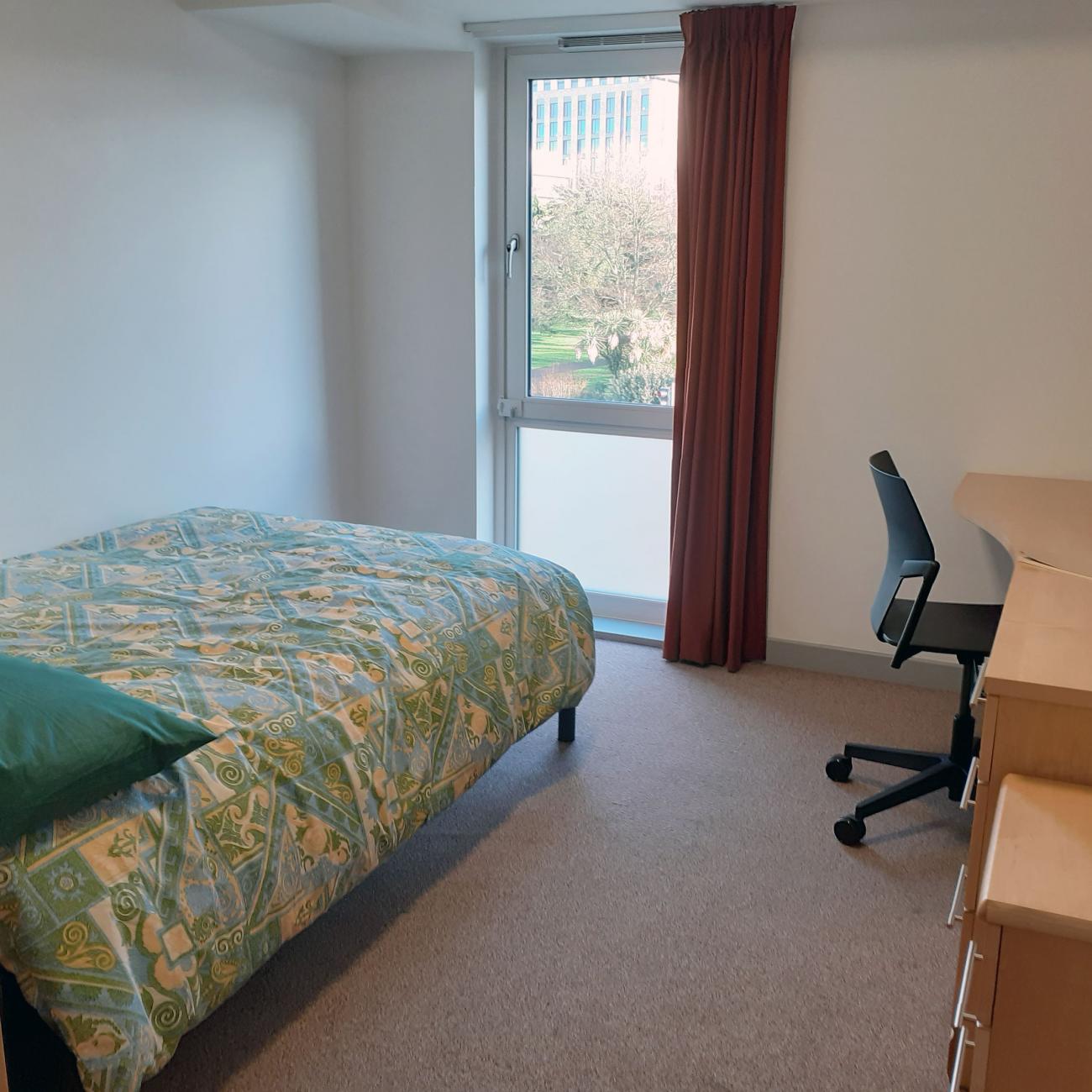 A student bedroom showing a double bed, large wooden desk and large window.