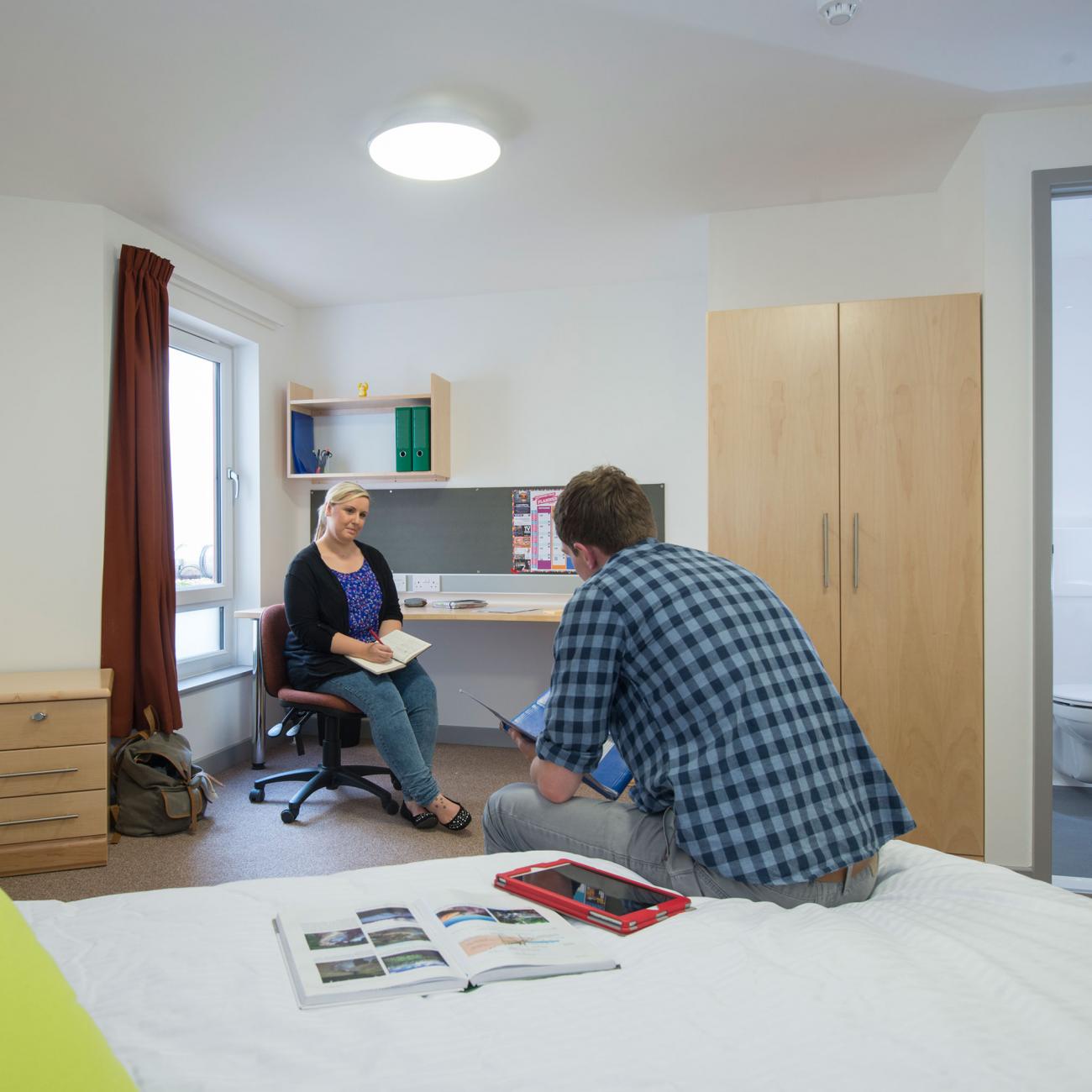 Two students sat talking in a large spacious bedroom. A double bed, desk and open door to a bathroom can be seen.