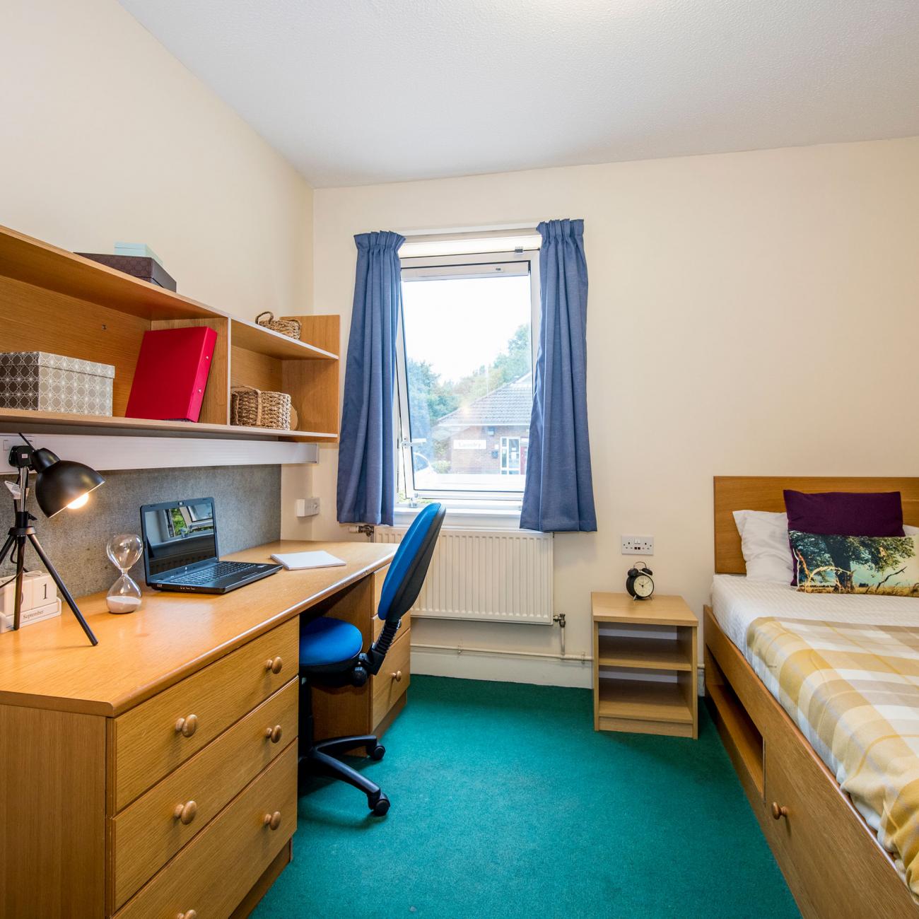 A student bedroom. A section of green carpet separates a single bed with patterned sheets and a large wooden desk. A window on the rear wall lets light in.
