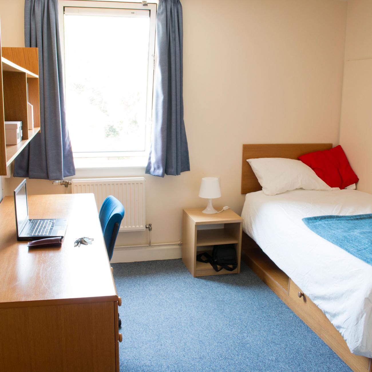 A student bedroom. A section of blue carpet separates a single bed with patterned sheets and a large wooden desk. A window on the rear wall lets light in.