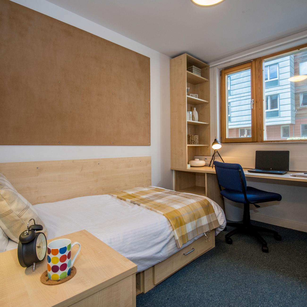 An empty student bedroom containing a neatly made single bed and a large desk with chair, beneath a window.