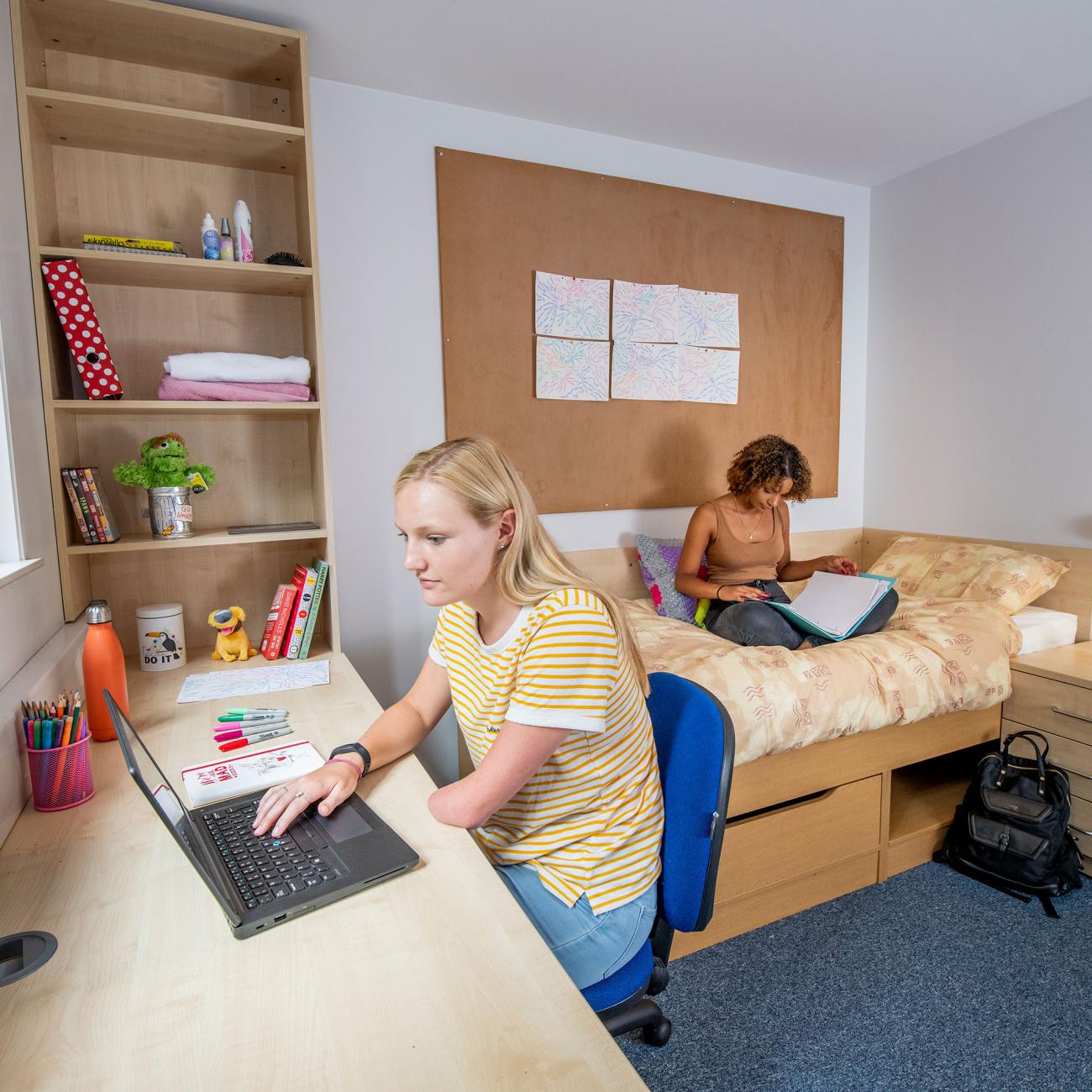 Two students in a bedroom. One sits reading on a single bed while the other works on a laptop, sat at a desk beside a window.