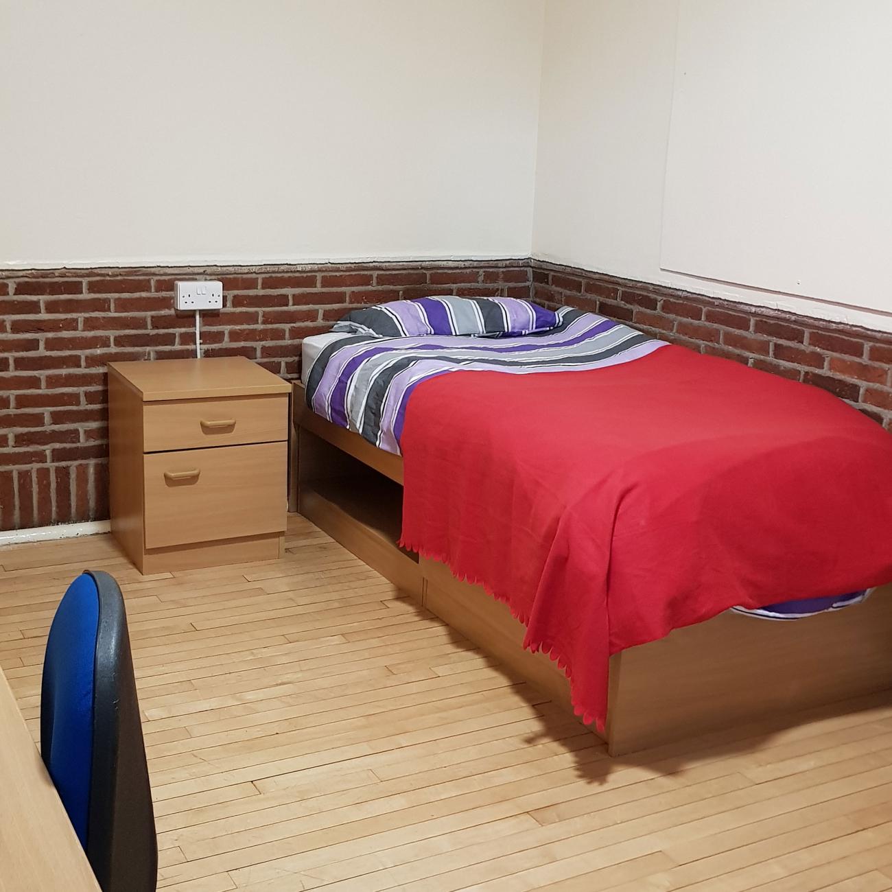 A single bed with red sheets in an empty student bedroom with wooden flooring.