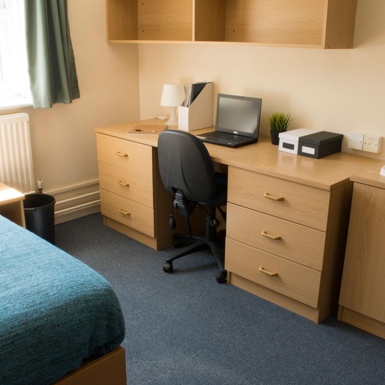 Student bedroom showing a desk and a neatly made bed with dark blue sheets. A large window lets light into the room.