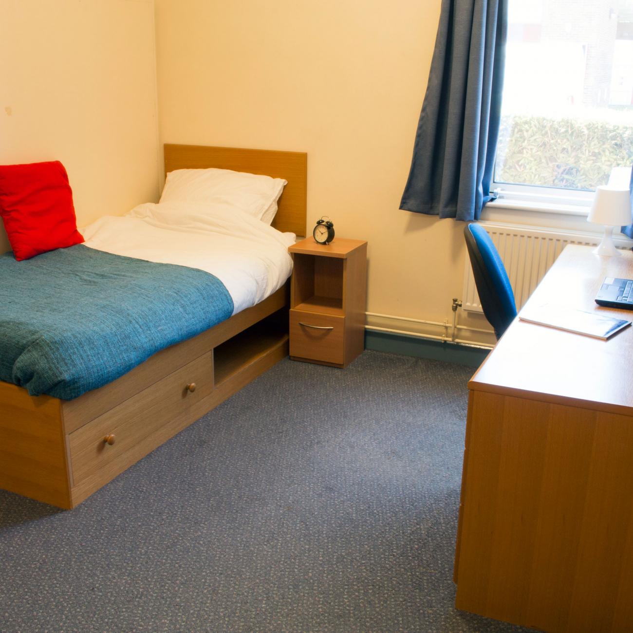 A spacious bedroom. The floor is blue carpeted and a neatly made single bed is visible against one wall. On the other side of the room is a wooden desk with office chair and a large window.
