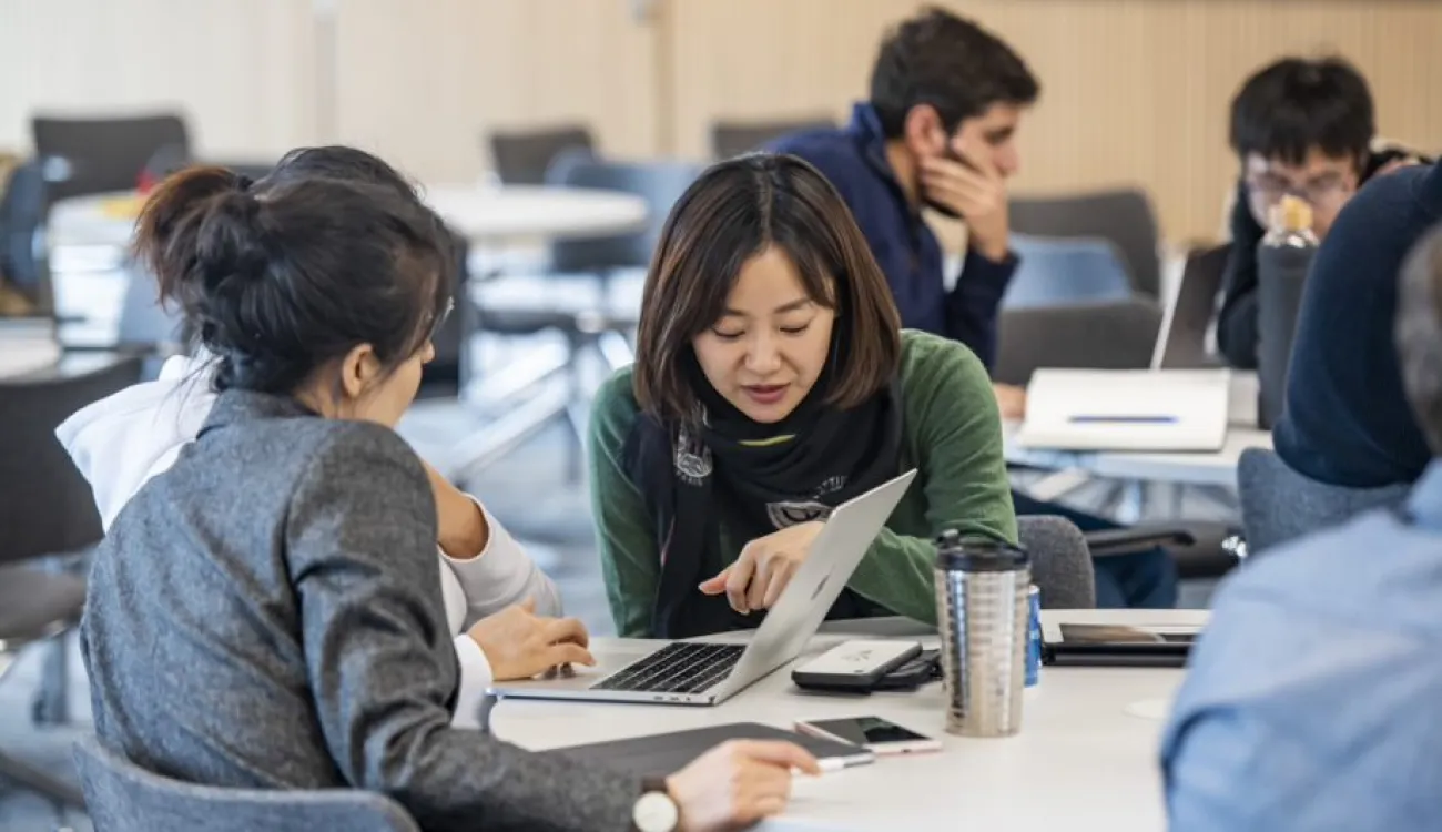 A small group of students sitting around a table look at a laptop screen and discuss their work.