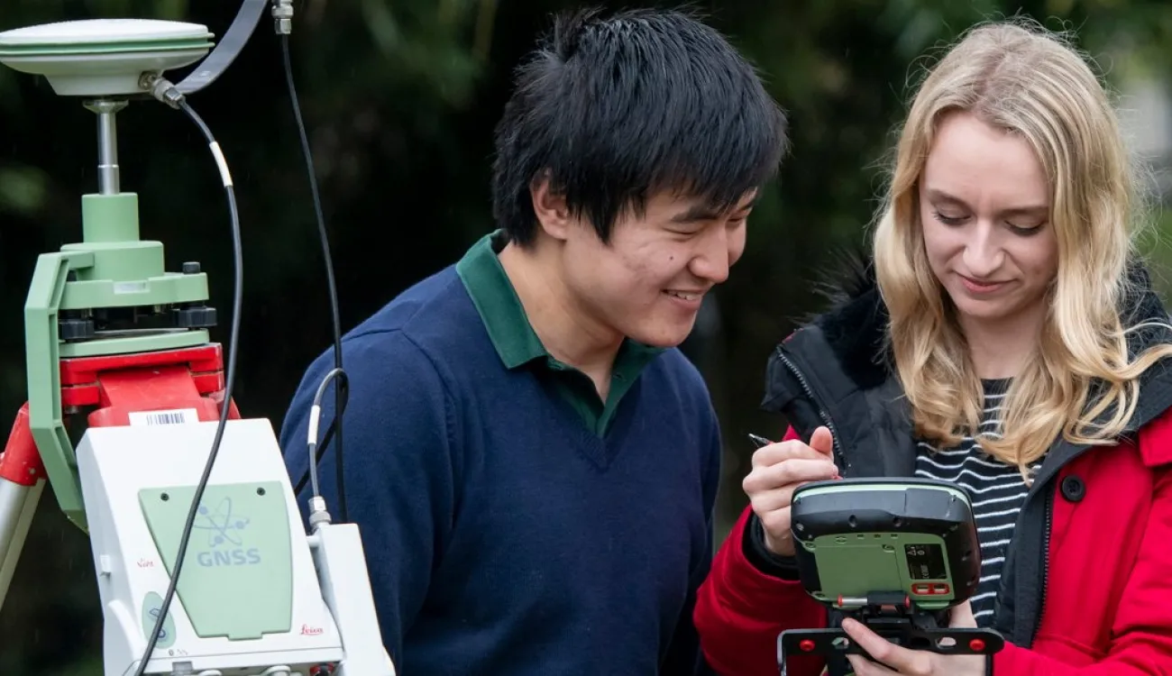 Two students working outdoors with field equipment. One is leaning over to look at the display that the other is studying.